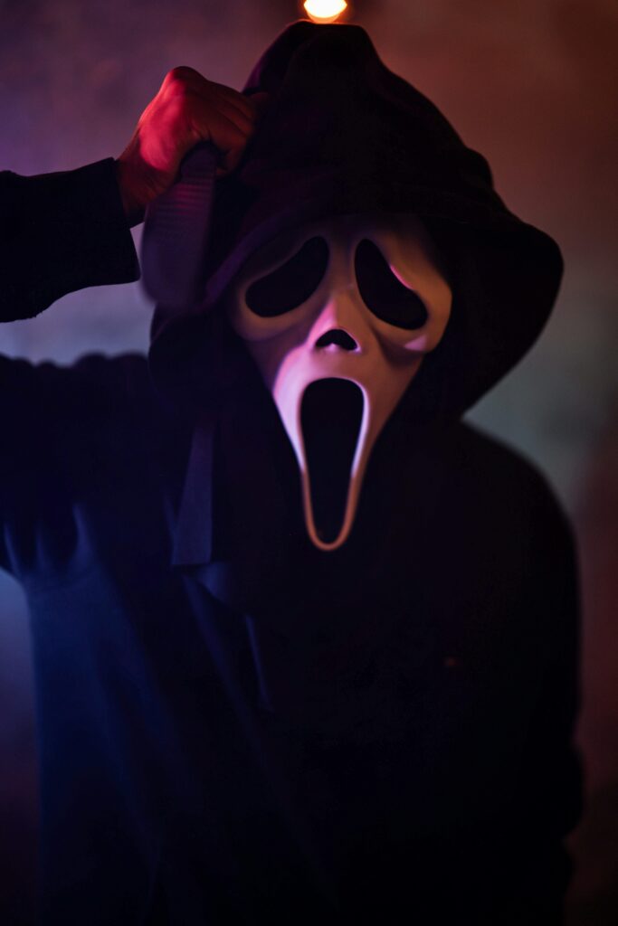 A menacing figure wearing a Halloween mask and hood in low light, creating an eerie atmosphere.